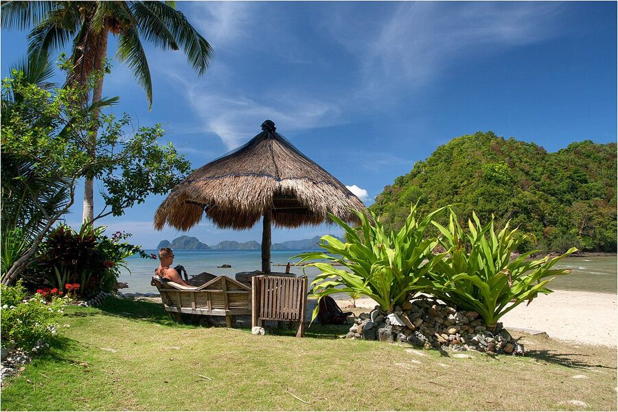 A thatched beach umbrella and lounger at Las Cabanas Beach, El Nido, with a view of forested headlands and turquoise water