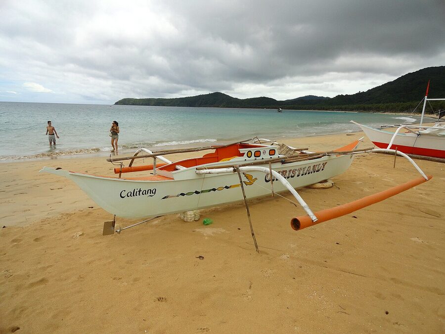 A bangka outrigger boat parked on the wide white sand of Nacpan Beach, El Nido, with two swimmers in the surf