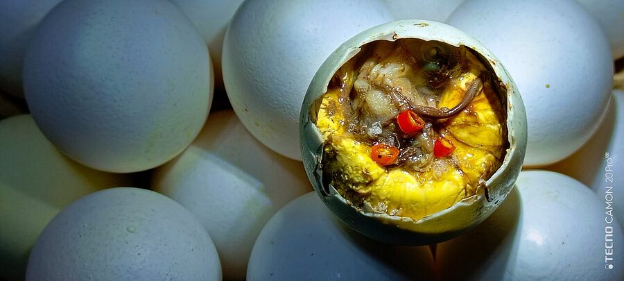 An open balut fertilised duck egg with chili and yolk visible inside the cracked shell beside other unopened balut eggs