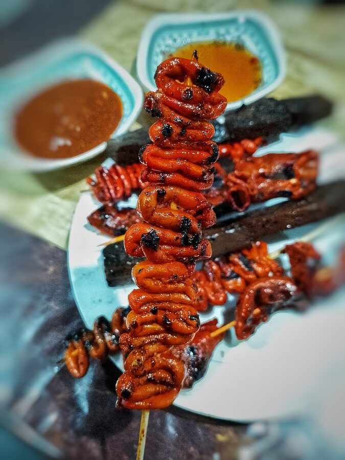 Grilled isaw chicken intestine skewers with smoky char and dipping sauces on a Manila ihaw-ihaw stand