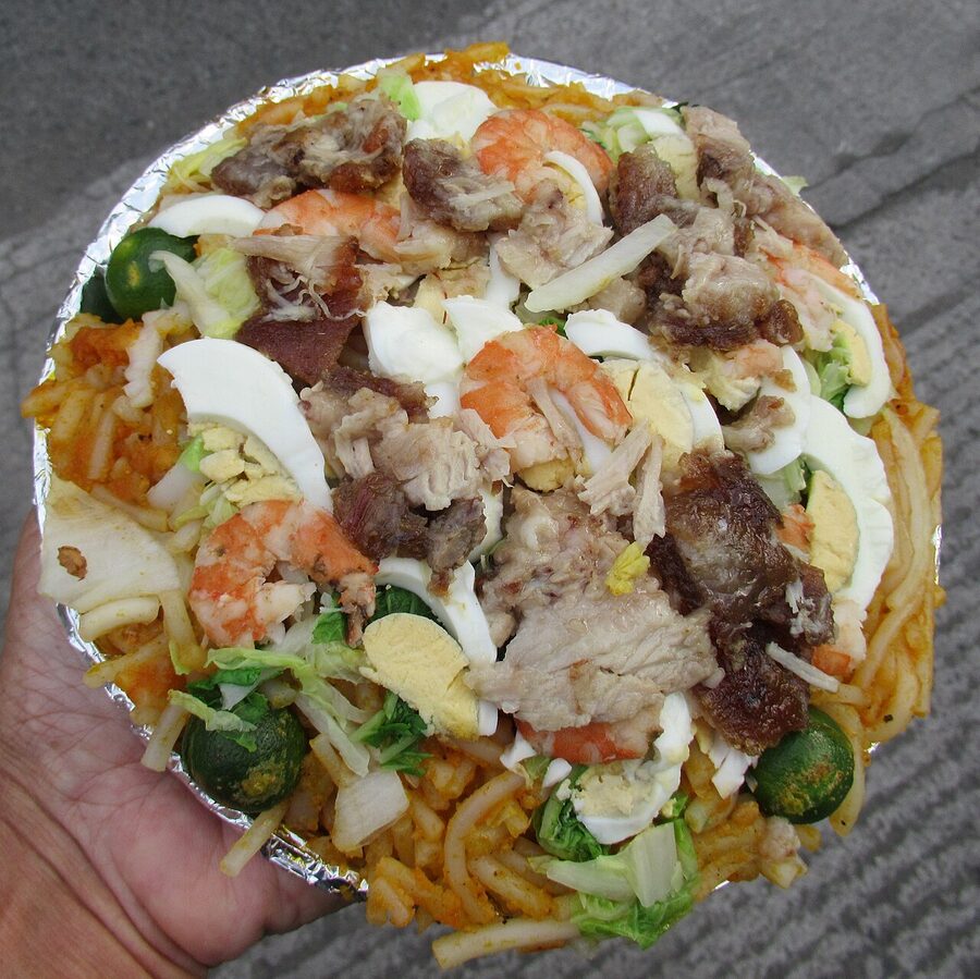 A hand holding a foil tray of pancit palabok with shrimp pork chicharon hard-boiled egg slices and calamansi